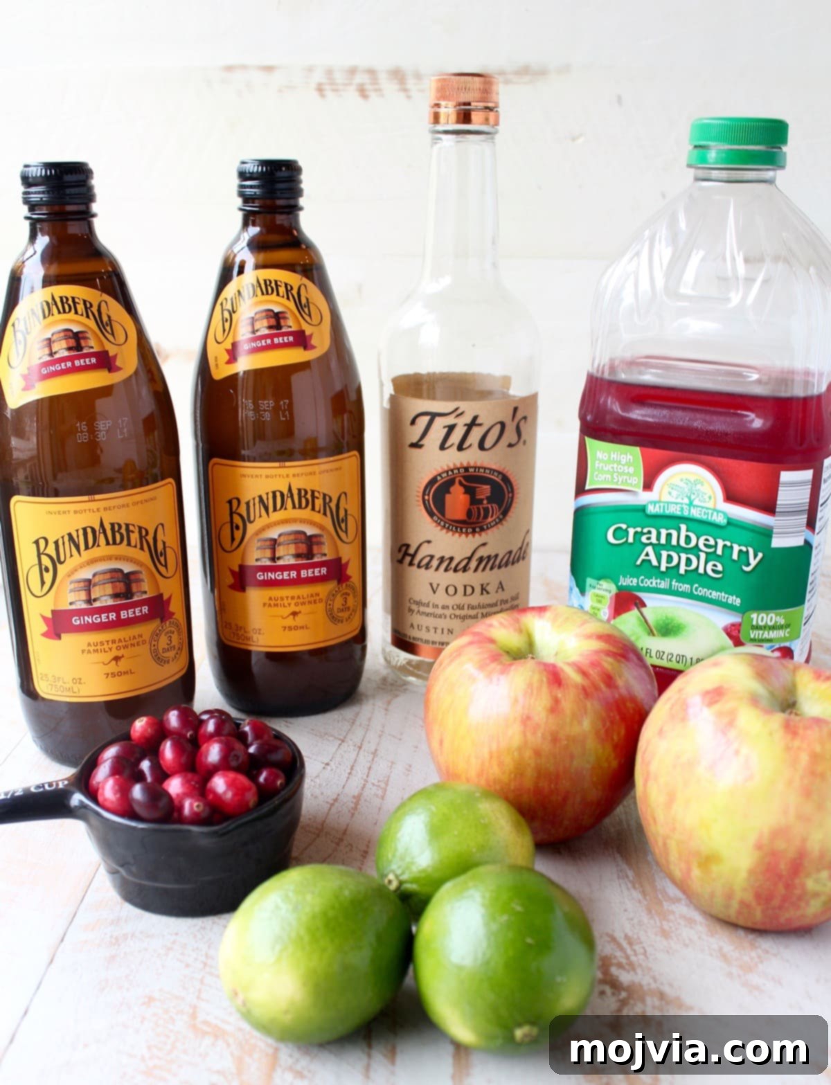 Cranapple Moscow Mule Punch 3 Ingredients for Cranberry Apple Moscow Mule Punch laid out on a white wooden board, including vodka, juices, ginger beer, cranberries, and apples.