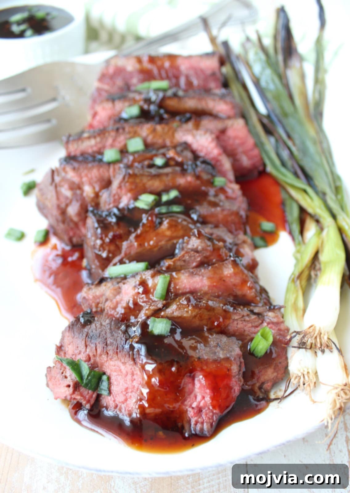 sliced cooked short ribs on a plate covered in Korean bbq sauce and diced green onions with grilled green onions on the side