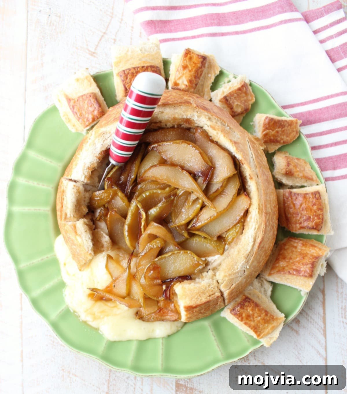 Decadent Caramelized Pear and Brie Bread Bowl 12 A beautifully presented Baked Brie Bread Bowl with caramelized pears and honey, served on a green plate with a festive red and white cheese spreader and a kitchen towel.
