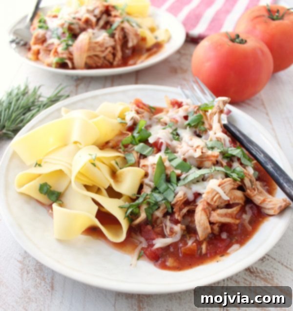 Tender Slow Cooker Chicken Ragu 2 Shredded chicken covered in red sauce, cheese, and herbs with pasta on a white plate.