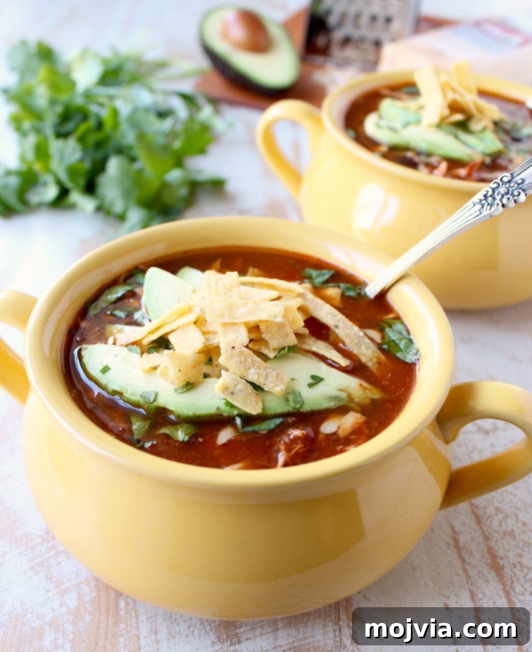 Creamy Chicken Enchilada Crockpot Feast 8 A vibrant yellow bowl of enchilada soup sits on a wooden table, adorned with perfectly sliced avocado and crunchy tortilla strips, with a second similar bowl softly blurred in the background, inviting a delicious meal.