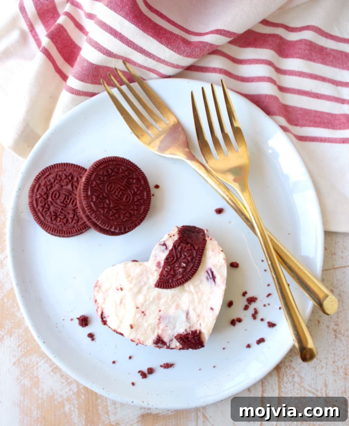 Heart-shaped Red Velvet Oreo cheesecake bar on a white plate with gold forks, showcasing the rich red crust and creamy filling.