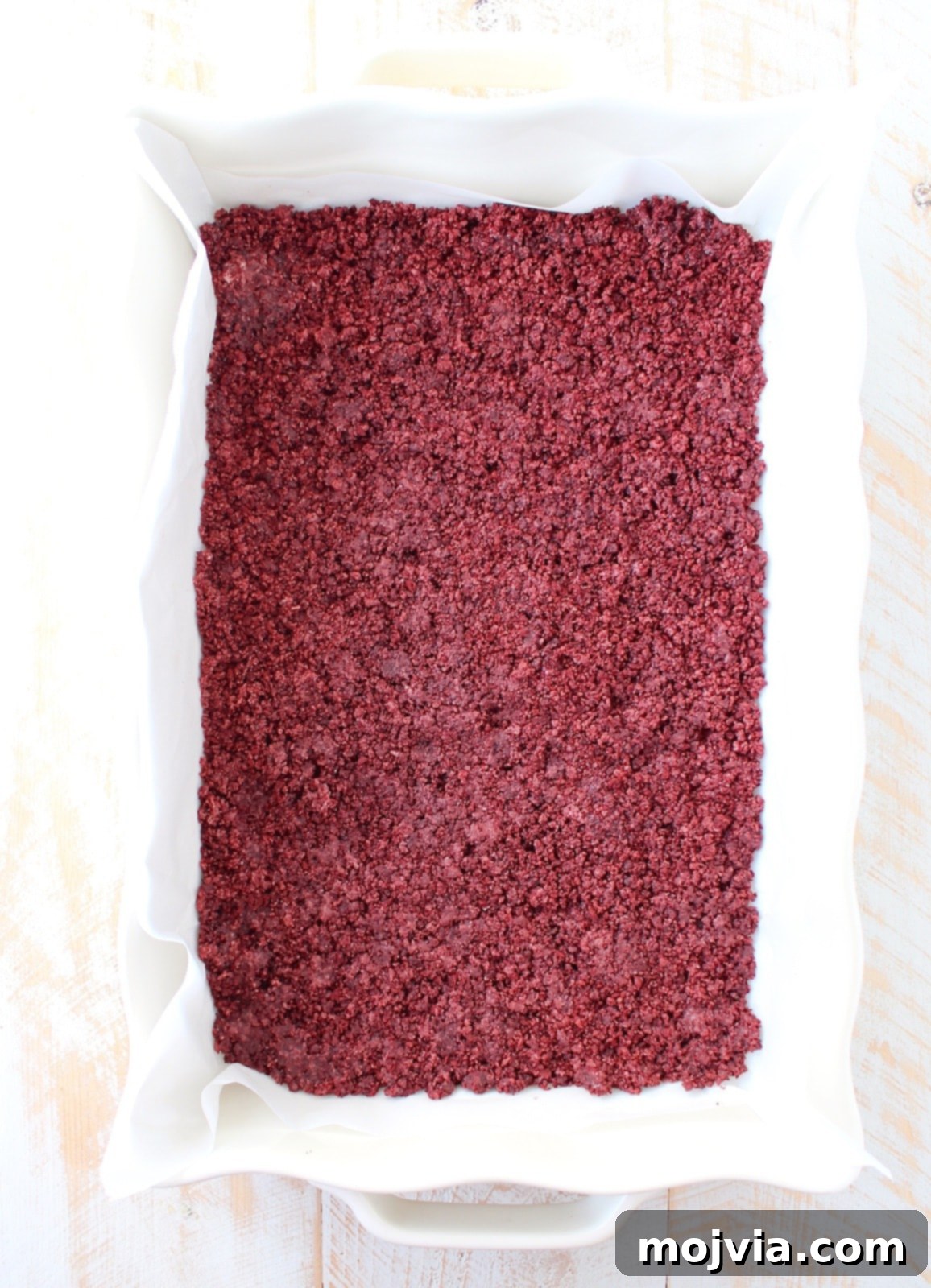 Finely crushed Red Velvet Oreos mixed with melted butter, pressed into the bottom of a parchment-lined baking dish, forming a vibrant red crust.