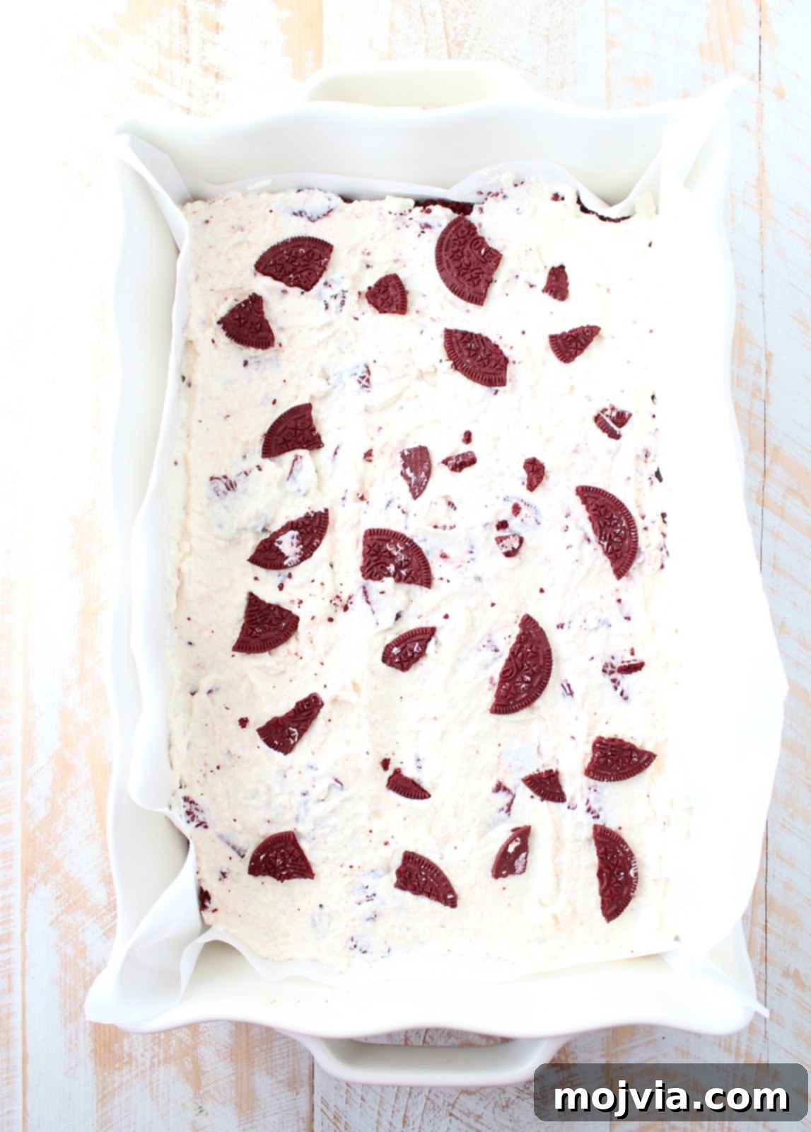 Assembled no-bake Red Velvet Oreo cheesecake bars in a baking dish, topped with additional cookie crumbles, ready for freezing.