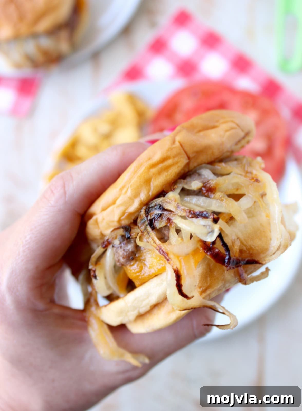 Oklahoma Onion Smash Burger 12 Hand holding a perfectly cooked Oklahoma Fried Onion Burger, showcasing its layers of beef, caramelized onions, and melted cheese, above a plate.
