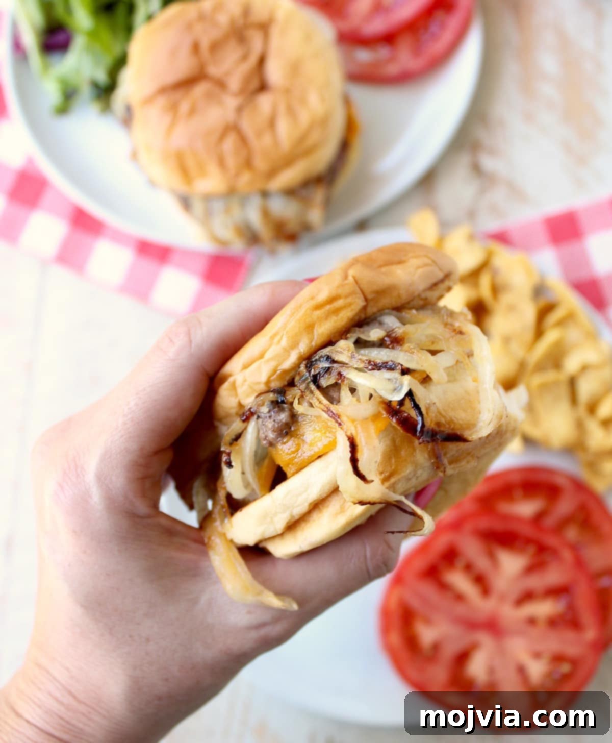 Oklahoma Onion Smash Burger 14 Close-up of a hand holding an Oklahoma Fried Onion Burger, ready to be enjoyed, with side dishes visible on a plate.