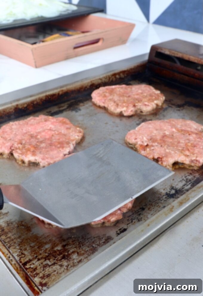Oklahoma Onion Smash Burger 7 Ground beef balls being smashed into thin burger patties on a hot griddle using a metal spatula.