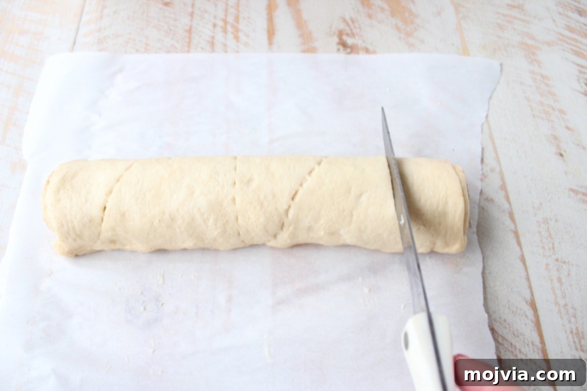 A chef's knife slicing a rolled crescent dough log into individual cinnamon roll spirals on parchment paper.