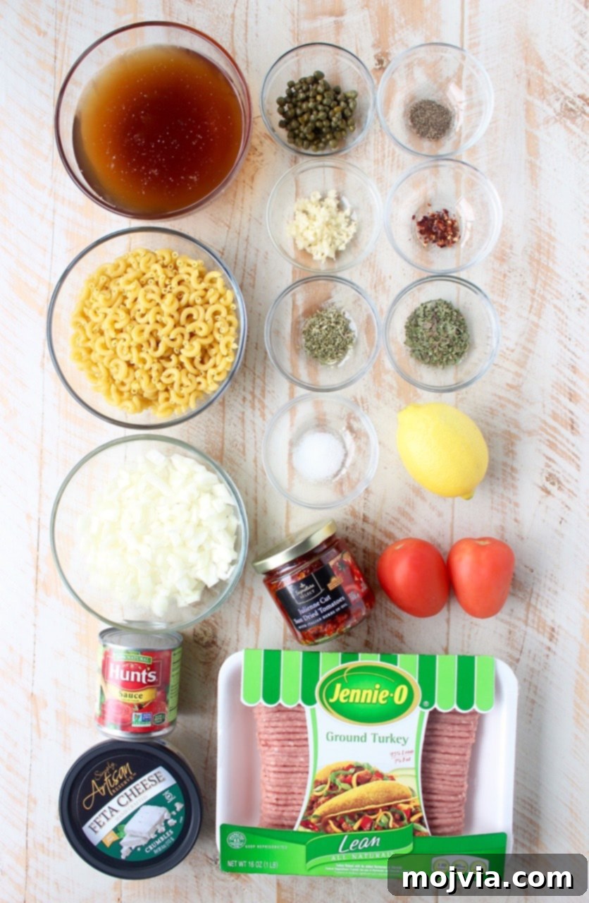 Fresh ingredients laid out on a white wooden board for one pot Greek pasta, including olive oil, onions, garlic, ground turkey, various spices, sun-dried tomatoes, Roma tomatoes, capers, tomato sauce, macaroni noodles, beef broth, lemon, and feta cheese.