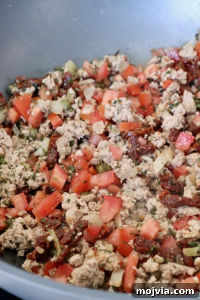 Ground turkey in a pan after being seasoned with herbs and spices, combined with diced tomatoes and capers, simmering gently.