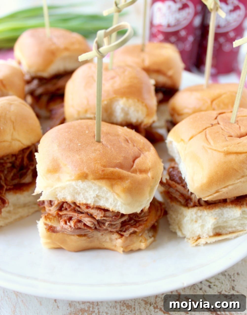 Sweet and Smoky Dr Pepper Pulled Pork Sliders 7 Mini pulled pork Hawaiian roll sandwiches on a white plate with a toothpick in each one for serving.