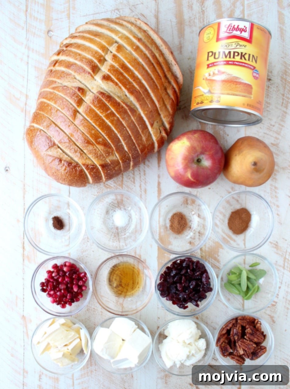 Pumpkin toast ingredients and various topping options artfully arranged on a rustic white wooden board. Ingredients include a can of pumpkin puree, a loaf of sourdough bread, and small bowls of ground cinnamon, nutmeg, cloves, and sea salt. Toppings feature fresh apple slices, pomegranate seeds, dried cranberries, candied pecans, a wedge of brie cheese, sliced pears, and fresh sage leaves.