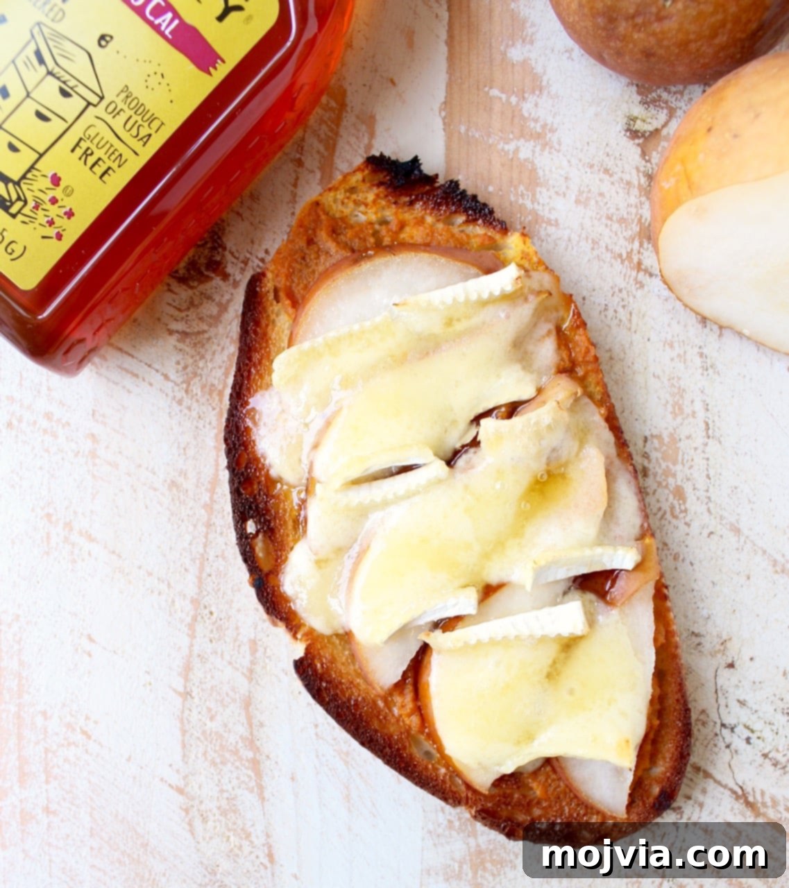 A delectable slice of pumpkin toast, generously topped with creamy melted brie cheese, delicate slices of fresh pear, and a glistening drizzle of golden honey, presented on a white wooden board.