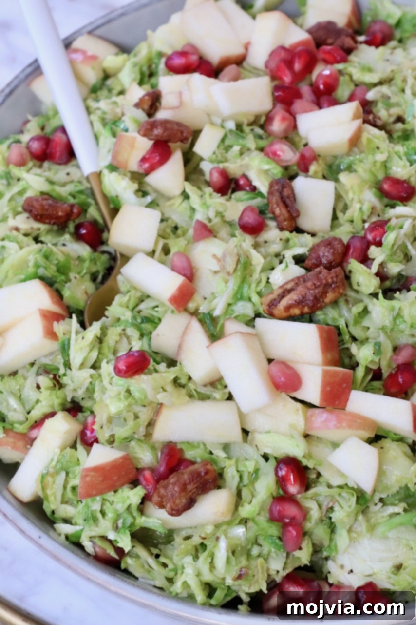 A large bowl of finished Brussels sprout salad, generously topped with diced apples and gleaming pomegranate seeds, ready to be served.