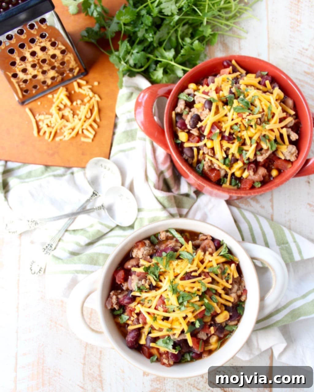 One-Pan Fiesta Turkey Soup 2 Two bowls filled with chili and shredded cheese on a wooden surface surrounded by a green and white napkin, spoons, herbs, and a cutting board with shredded cheese.