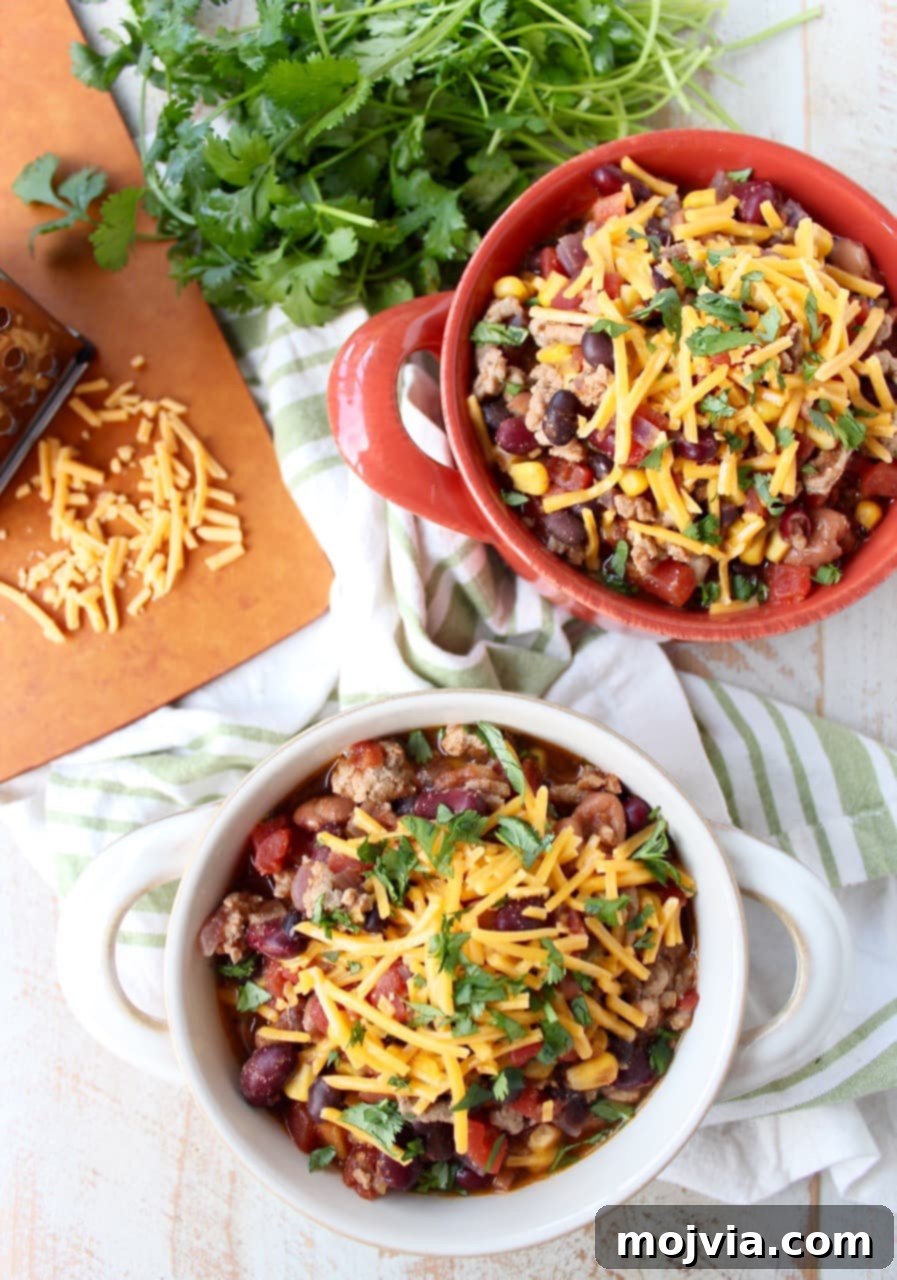 One-Pan Fiesta Turkey Soup 5 Two bowls filled with chili and shredded cheese on a wooden surface surrounded by a green and white napkin, spoons, herbs, and a cutting board with shredded cheese.
