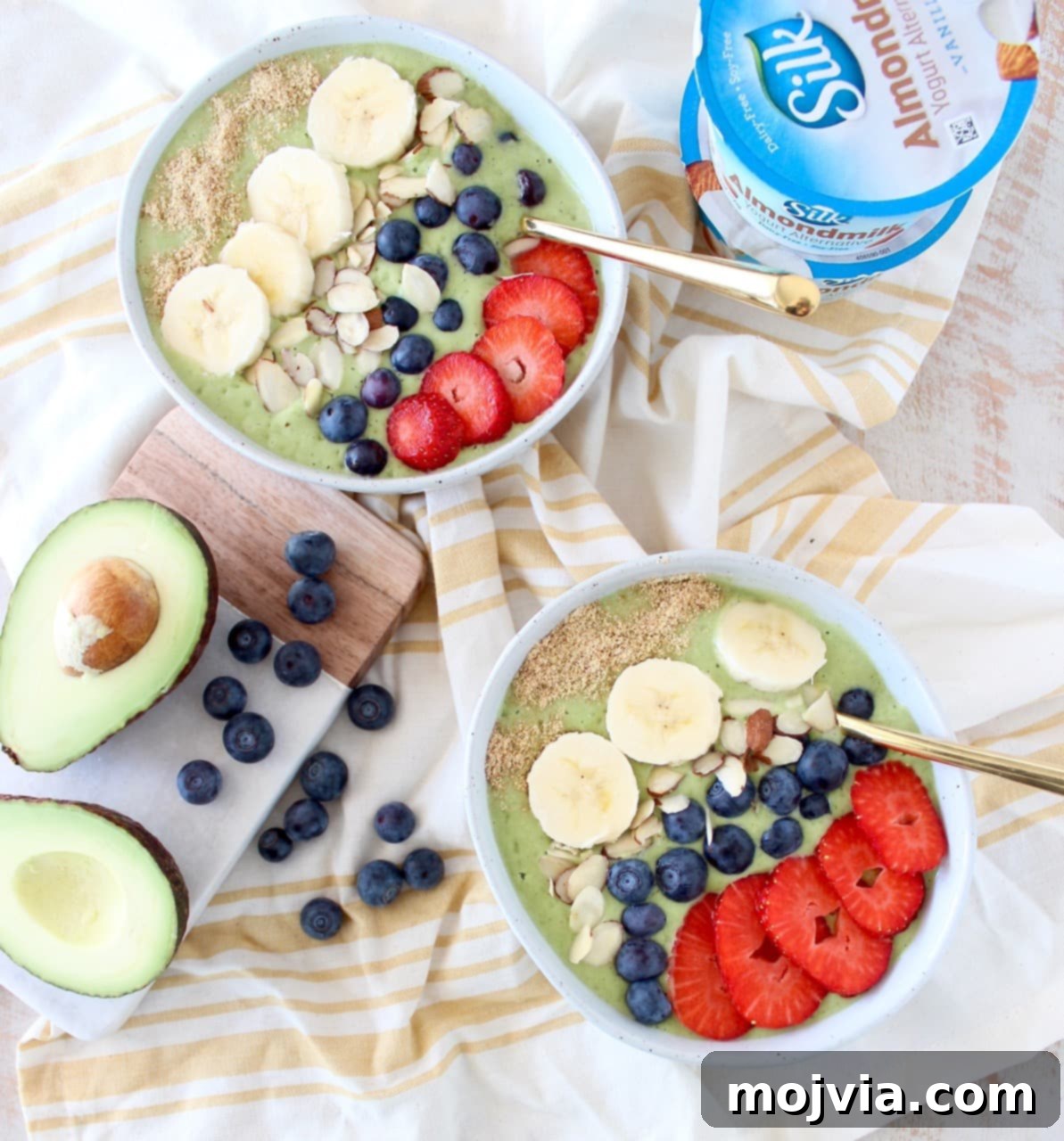 Two vibrant green avocado smoothie bowls topped with an array of fresh fruit and nuts, including sliced banana, blueberries, and almonds.