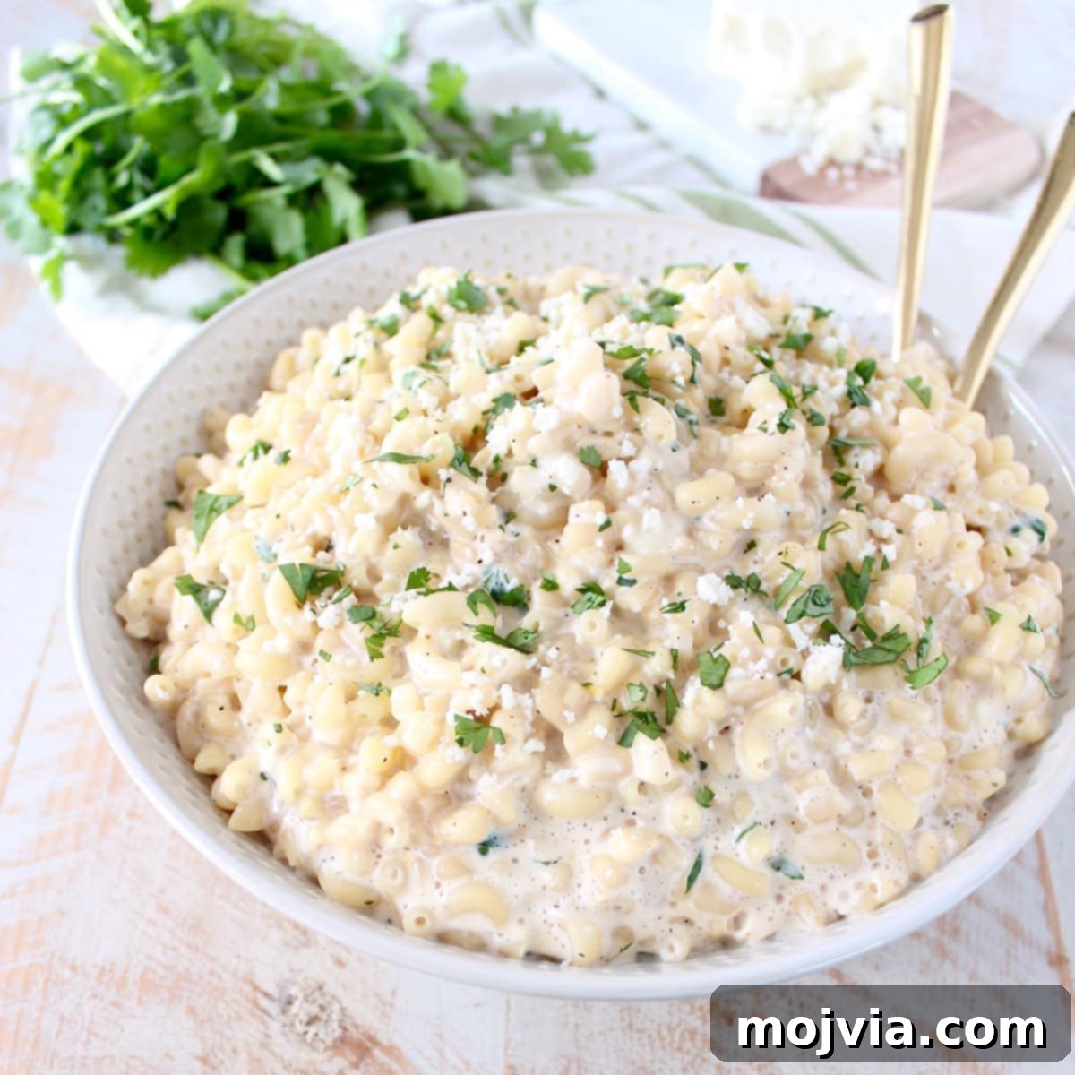Street Corn Fiesta Mac 3 A white bowl of Mexican street corn mac and cheese with gold serving spoons nestled in, highlighting its creamy texture and rich color. The dish is topped with fresh green herbs.