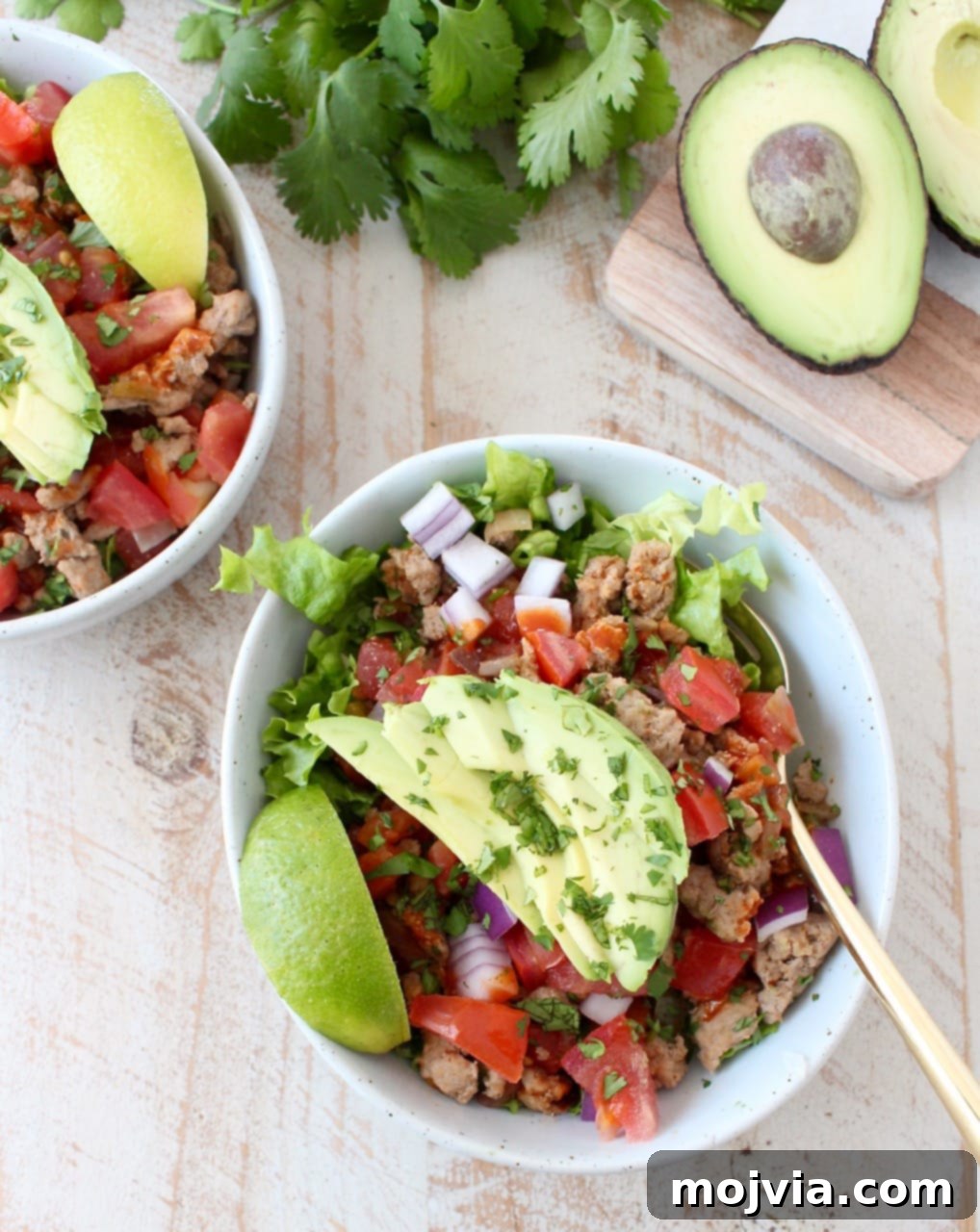 Whole30 Power Turkey Taco Salad 3 A rustic wooden surface holds a white bowl generously filled with vibrant Taco seasoned ground turkey, peppers, onions, and tomatoes, all nestled on a bed of crisp lettuce.