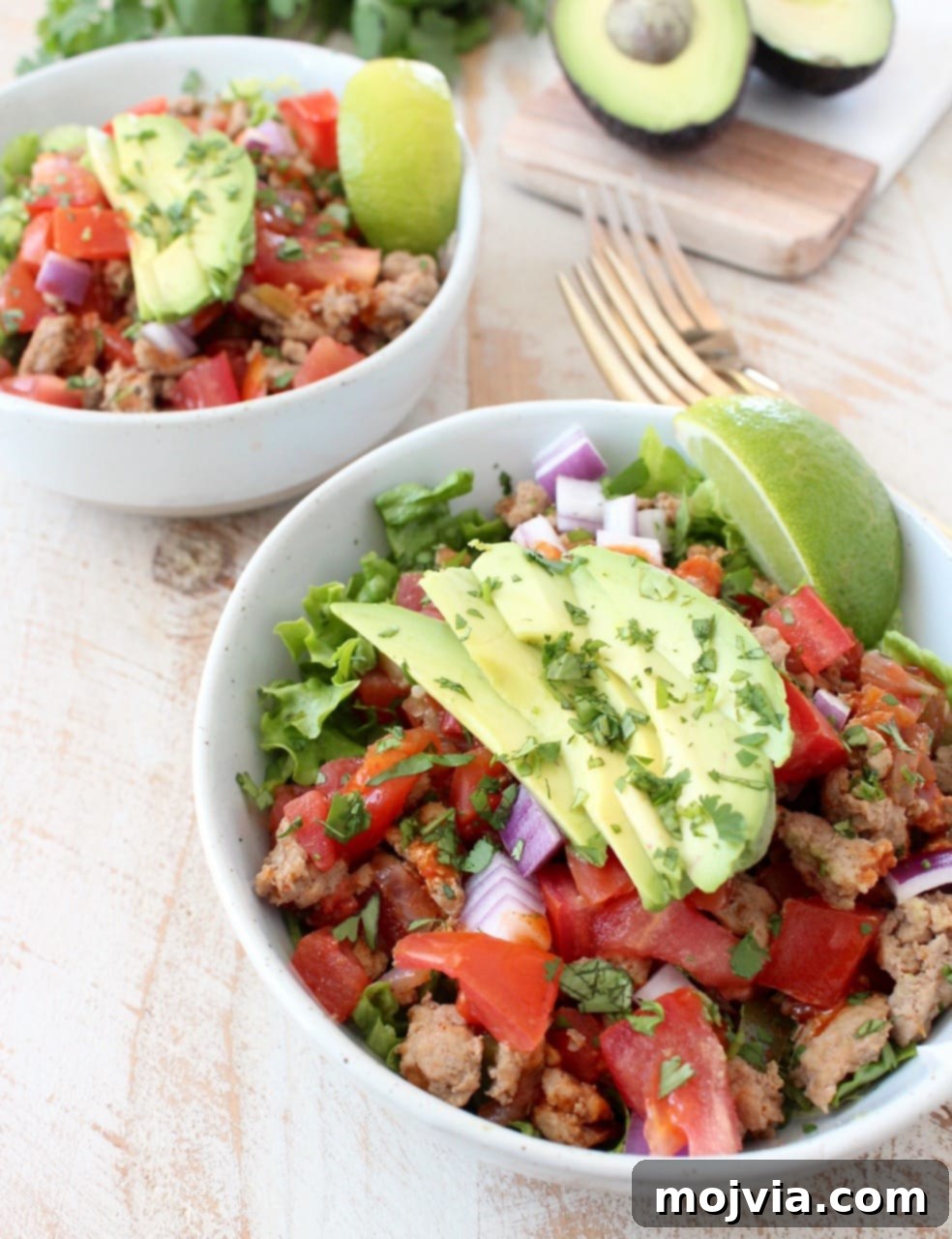 Whole30 Power Turkey Taco Salad 5 A side view of two inviting white bowls, each brimming with taco seasoned ground turkey cooked with peppers, onions, and tomatoes, adorned with fresh avocado slices, all resting on a wooden surface.