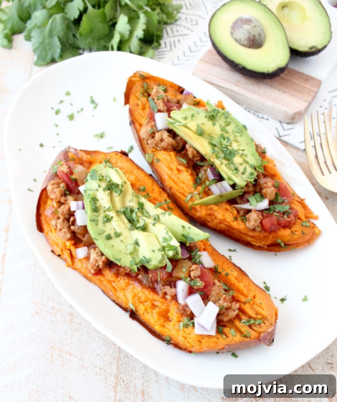 Baked sweet potato sliced in half, generously topped with chipotle chili, fresh avocado slices, and chopped cilantro on a white plate, ready to be enjoyed.