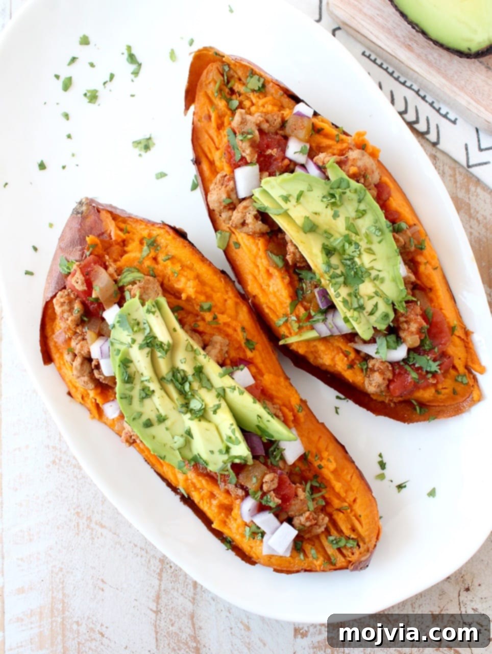 Another view of a baked sweet potato sliced in half, generously topped with chipotle chili, fresh avocado slices, and chopped cilantro on a white plate.