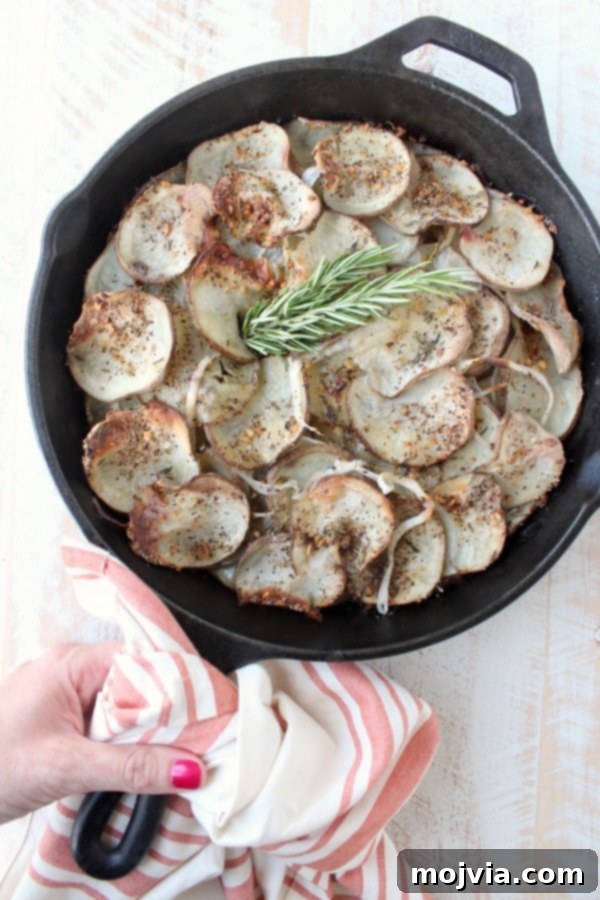 A hand holding a cast iron skillet over a wooden surface, showcasing the beautifully layered and seasoned potatoes ready for baking.
