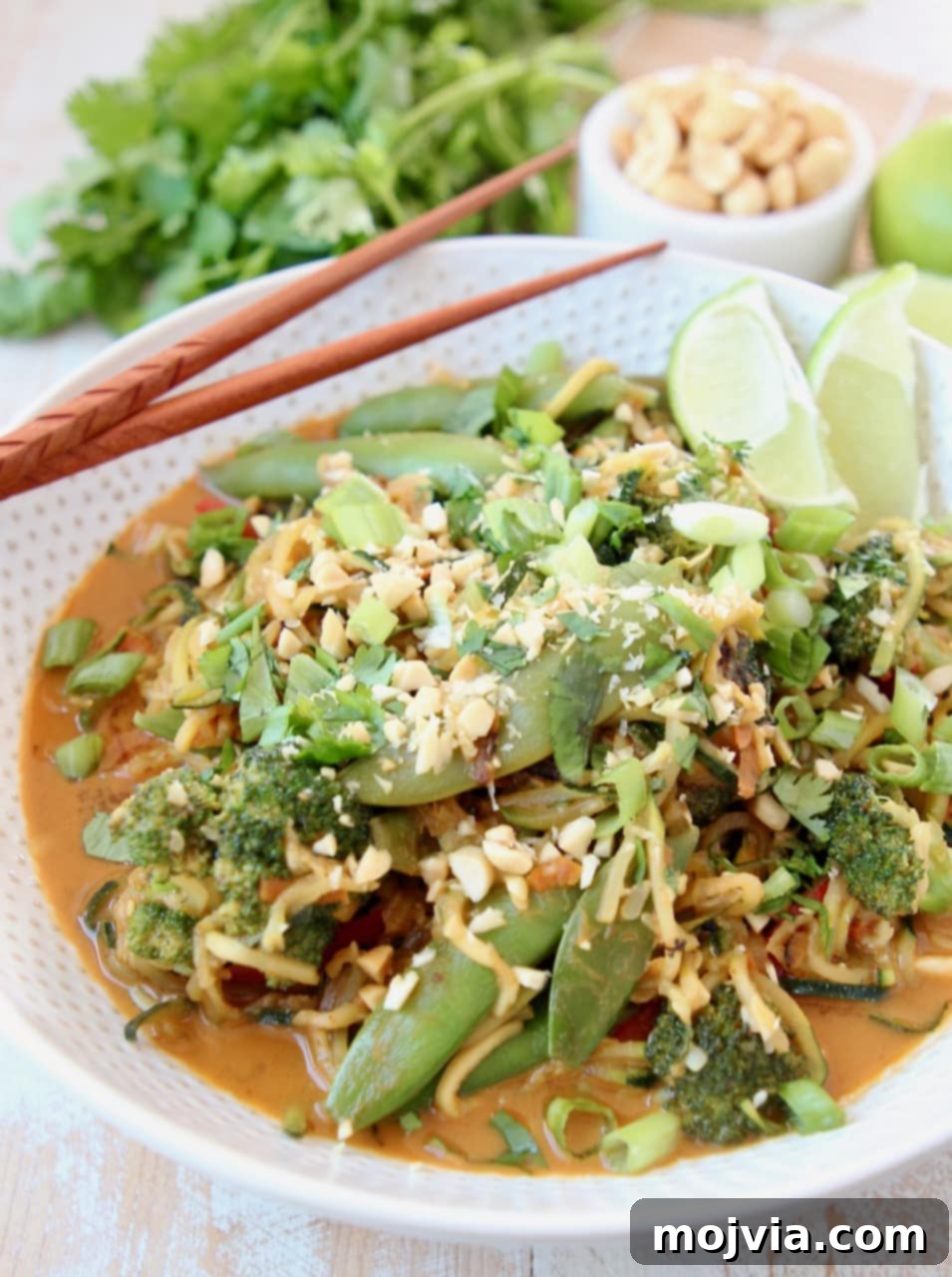 Zucchini Noodle Pad Thai: A Fresh Vegan Spin 3 A close-up shot of Vegan Pad Thai with Zucchini Noodles, featuring vibrant broccoli florets, crisp sugar snap peas, shredded carrots, all coated in a rich peanut sauce and garnished with fresh scallions and lime wedges in a rustic bowl.