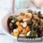 Baked gnocchi, butternut squash and kale in a bowl with wooden serving spoons, with a whole butternut squash and green baking dish in the background.