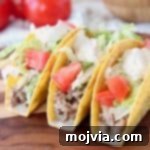 Caramelized Onion Taco Bliss 2 Ground beef tacos in crispy shells with diced tomatoes and shredded lettuce on a wooden cutting board with whole tomatoes in the background