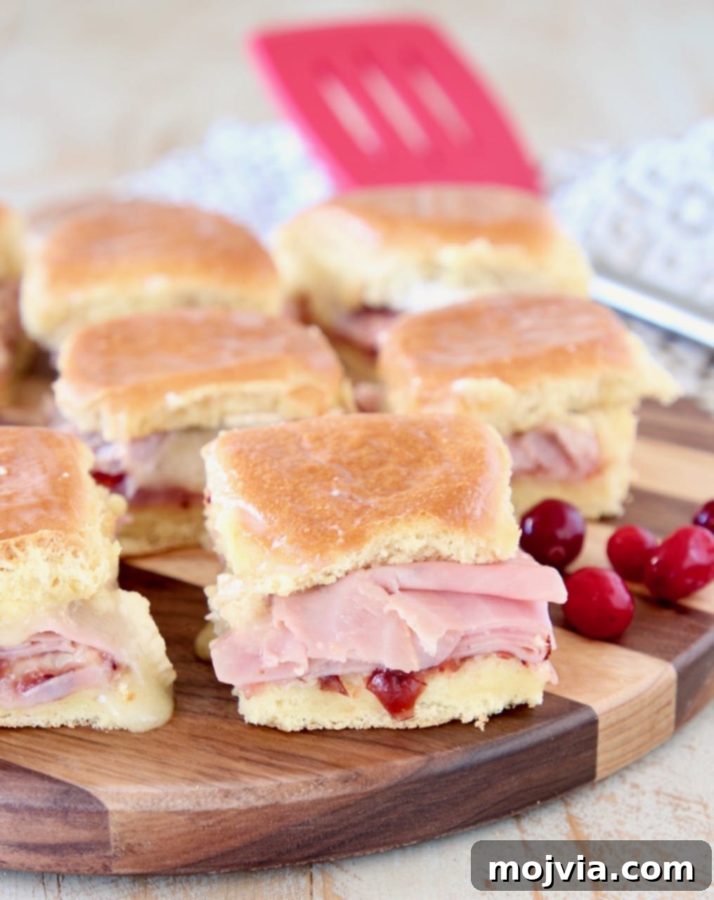 Sweet & Savory Cranberry Brie Ham Sliders 2 Golden brown Cranberry Brie Ham Sliders on Hawaiian rolls with fresh cranberries and a red spatula in the background, presented on a rustic wood cutting board.
