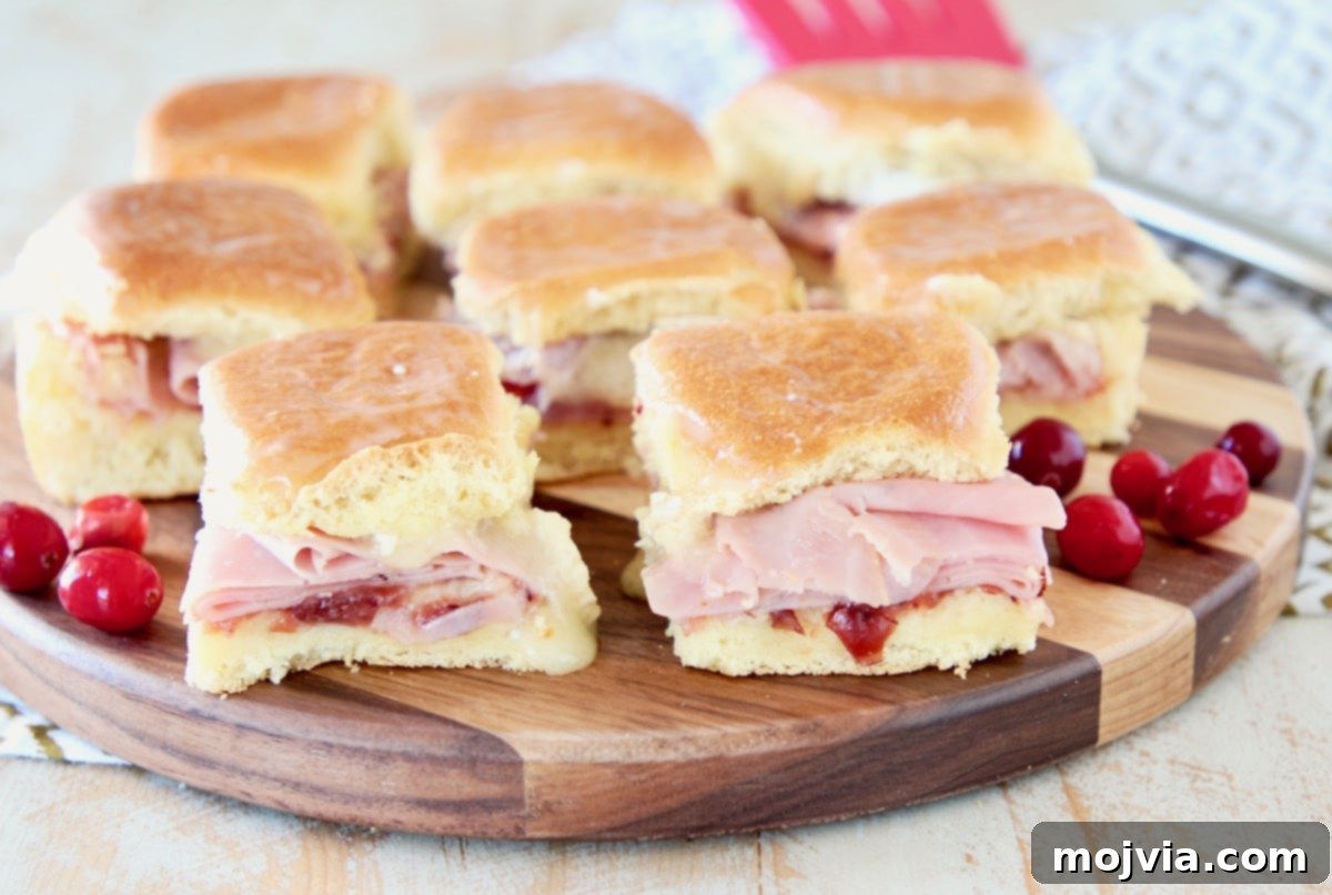 Sweet & Savory Cranberry Brie Ham Sliders 4 Close-up of baked Cranberry Brie Ham Sliders, showcasing the melted brie cheese and cranberry sauce, on a wooden board with a red spatula in the background.