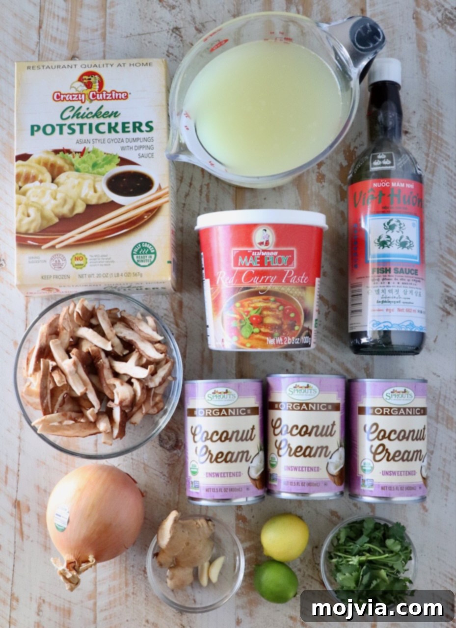 A beautiful flat lay of fresh ingredients for Thai coconut potsticker soup, including whole garlic cloves, a knob of ginger, stalks of lemongrass, red curry paste, cans of coconut milk, shiitake mushrooms, an onion, and a bag of frozen potstickers, all arranged on a rustic white wooden board.