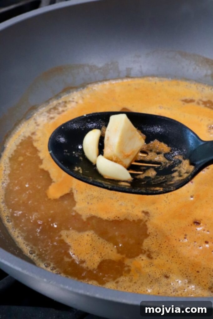 A slotted spoon carefully holding removed garlic cloves and a piece of ginger, lifted above a steaming pot of aromatic broth, indicating the next step in preparing the Thai coconut soup.