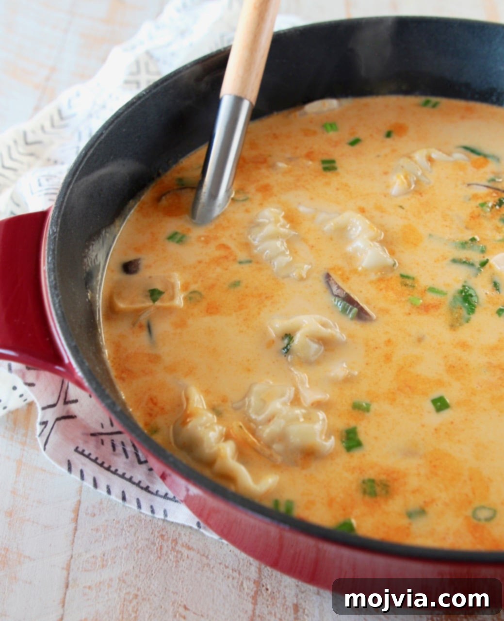 A vibrant red cast iron pot brimming with Thai coconut soup and potstickers, beautifully garnished with fresh cilantro and vibrant green scallions. A wooden spoon handle peeks out from the delicious soup.