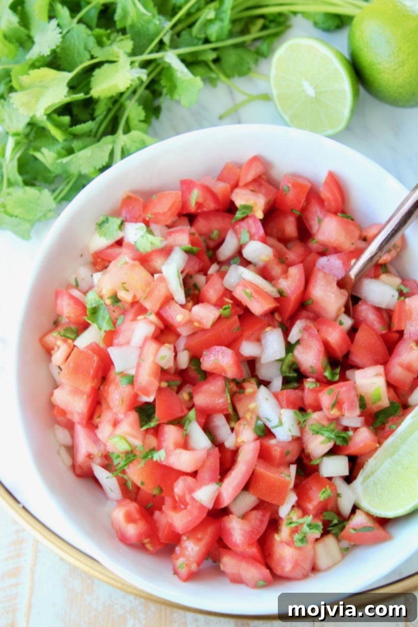 Fiesta Fresh Salsa 7 Overhead shot of pico de gallo in white bowl with fresh lime wedges and spoon in the bowl, with fresh cilantro on the side