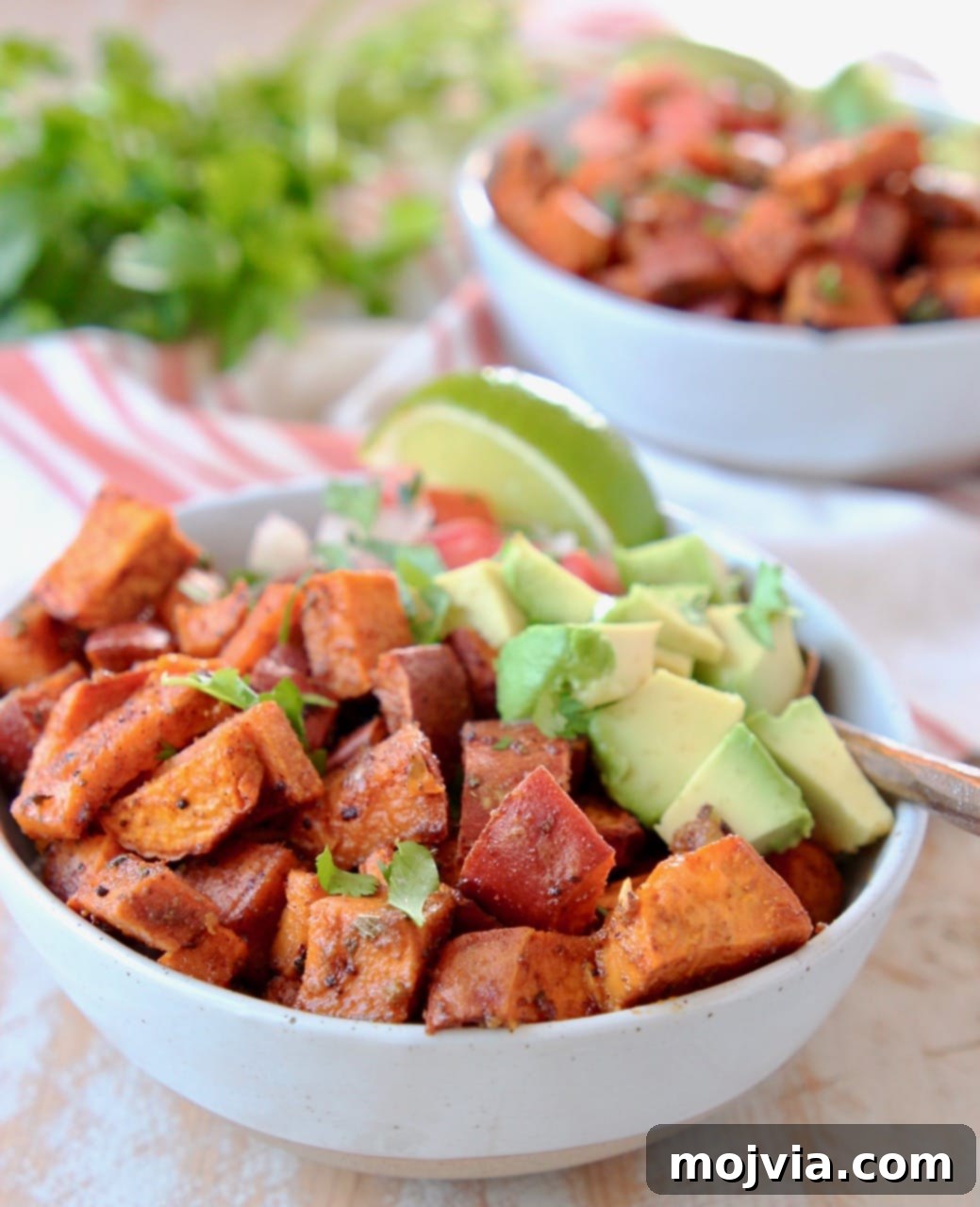 Southwest Vegan Sweet Potato Quinoa Fiesta 3 Diced roasted sweet potatoes and diced avocado in quinoa bowl with lime wedge and fork, with orange and white striped towel underneath and cilantro in background