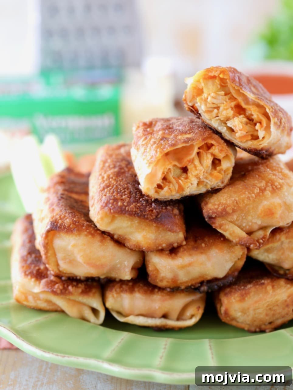 Crispy pan-fried Buffalo chicken egg rolls piled high on a vibrant green plate, with one egg roll expertly sliced in half revealing its savory, cheesy filling. A block of sharp cheddar cheese and a grater sit in the background, hinting at the fresh ingredients.
