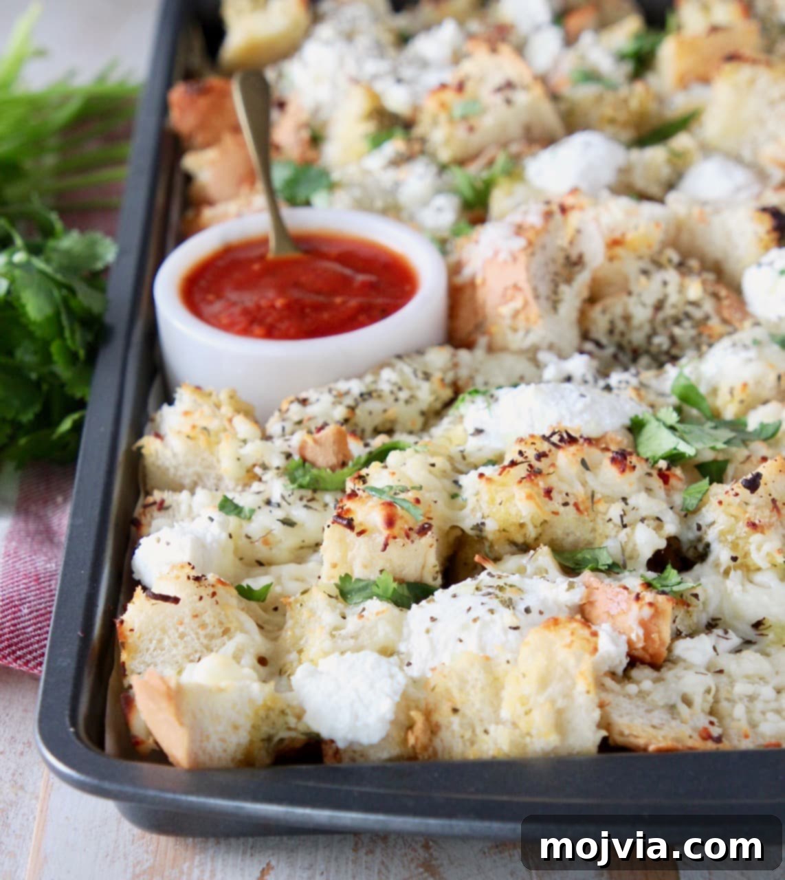 Cubed garlic bread, covered in cheese and fresh parsley on a baking sheet with a small bowl of marinara sauce for dipping