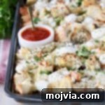 Cubed garlic bread covered in cheese on a baking sheet with a side of marinara sauce in a white bowl with a small spoon
