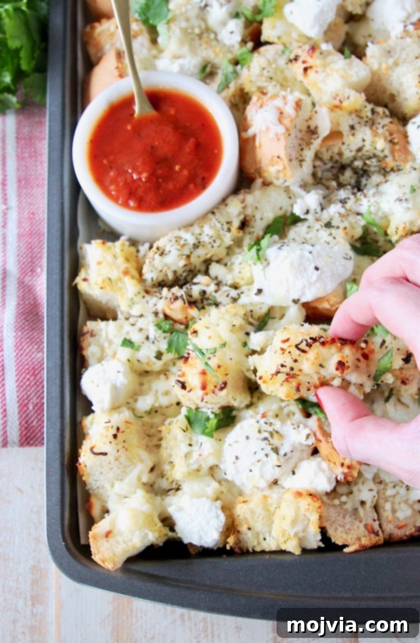 A hand reaching for a piece of cheesy garlic bread nacho from a baking sheet, with a small bowl of marinara sauce nearby.