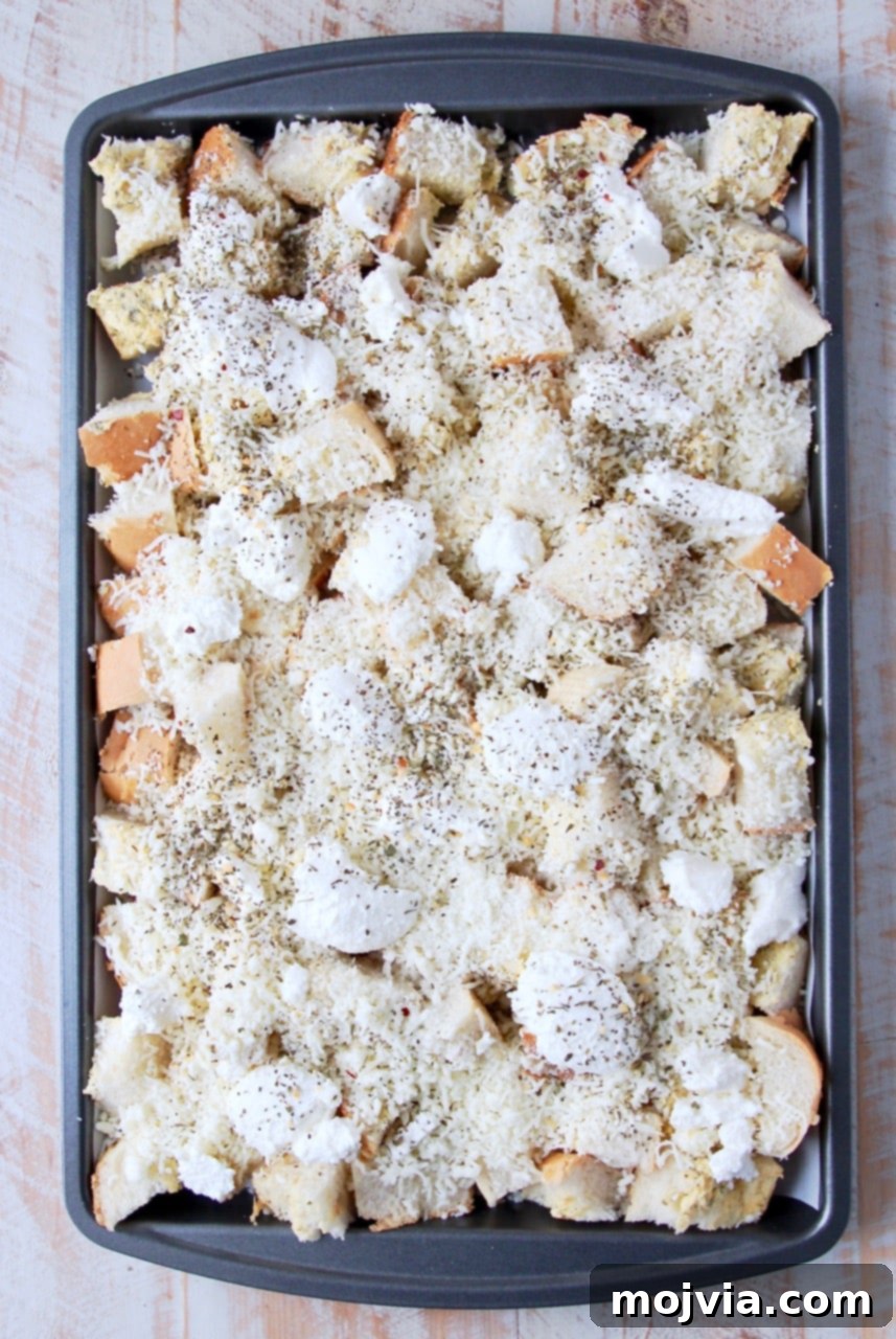 Garlic bread cubes generously covered in a variety of melted cheeses on a baking sheet, ready for baking.