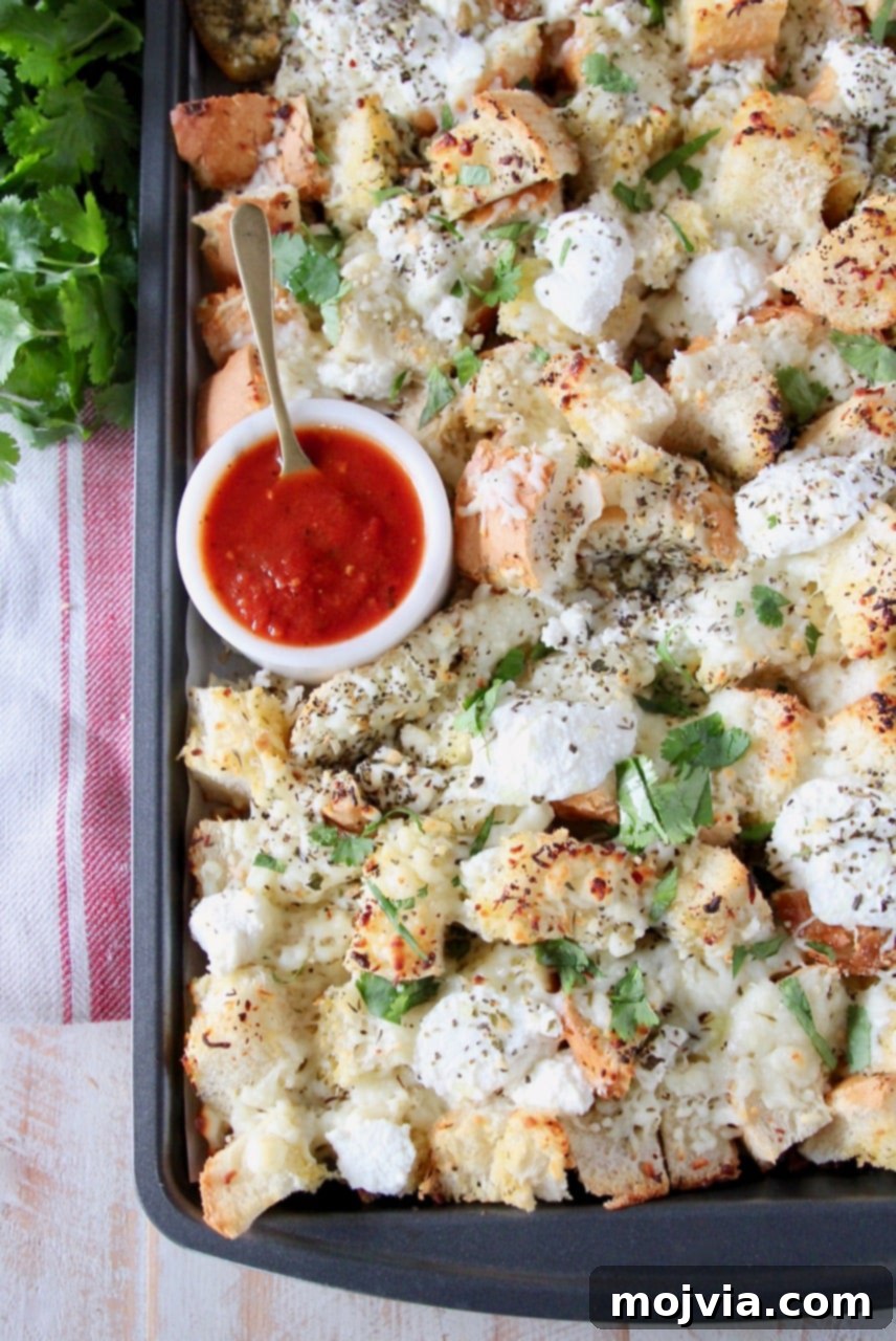 Close-up of cheesy garlic bread nachos on a baking sheet with a small bowl of marinara sauce and a spoon in the background.