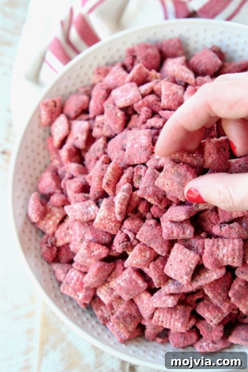 Crimson Velvet Crunch 3 Hand reaching into a white bowl to grab a piece of red velvet puppy chow, emphasizing its irresistible appeal