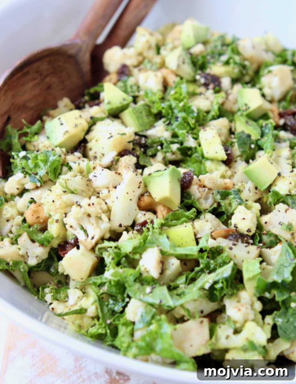 Cauliflower salad with kale, raisins and avocado in white bowl with brown wooden spoon