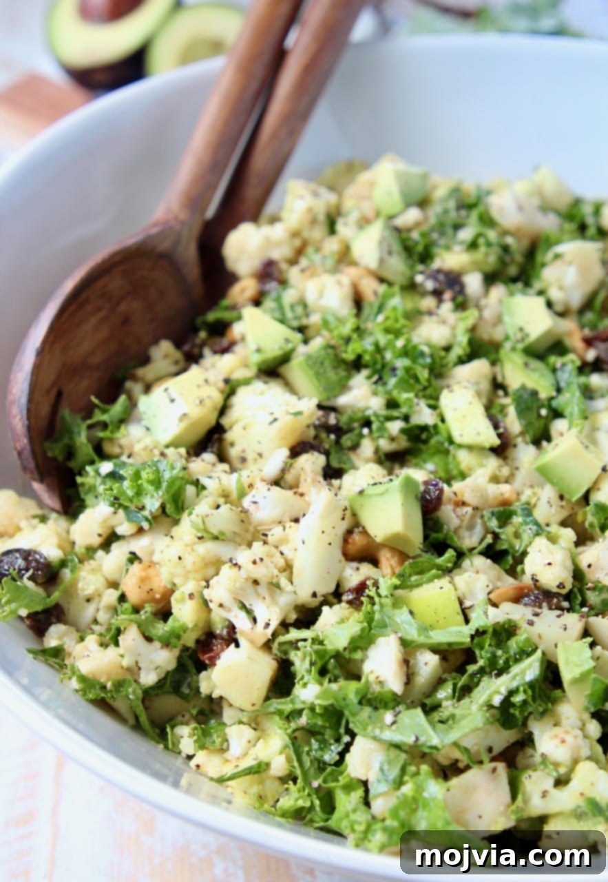 Diced cauliflower, kale, avocado and apples in large white bowl with wood serving spoons