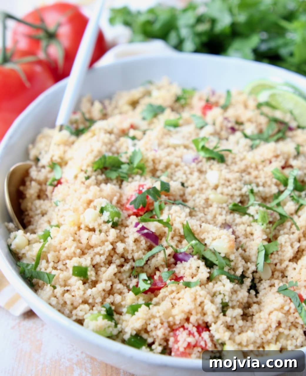 A vibrant bowl of Mexican Vegetarian Couscous, garnished with diced tomatoes, fresh cilantro, and lime wedges, showcasing the inviting textures and colors of roasted tomatillos and grilled corn.