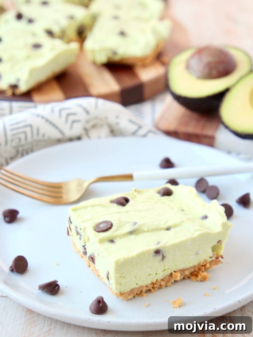 Effortless Avocado Bliss Bars 2 Avocado cheesecake bar with graham cracker crust on white plate with gold fork and chocolate chips. A refreshing and easy summer dessert idea.
