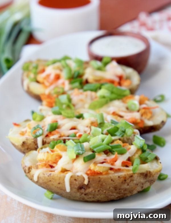 Set It and Forget It Buffalo Chicken 9 Buffalo chicken potato skins lined up on a white oval plate topped with diced green onions with a small bowl of ranch dressing in the background