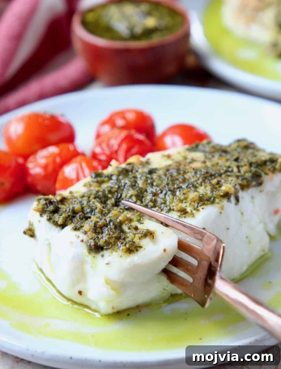 Pesto-Crusted Chilean Sea Bass 2 Fork cutting into flaky, perfectly cooked Chilean sea bass on a white plate, generously topped with vibrant green pesto and surrounded by roasted cherry tomatoes. The fish displays a beautiful golden-brown sear, promising a tender interior.